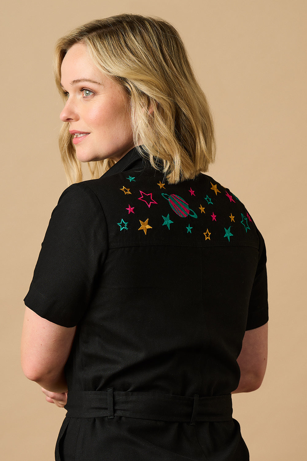 A black boilersuit with outer space-themed embroidery, featuring a planet and stars on the shoulders. The garment has a half sleeve shirt upper, an exposed front zip, and straight legs.
