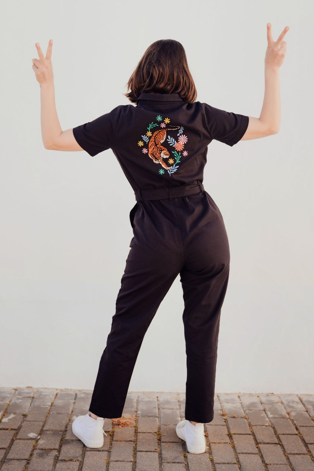 A person standing with their back towards the camera, wearing a black boilersuit with a tiger and floral embroidery on the back. They are making a peace sign with their hands.