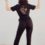 A person standing with their back towards the camera, wearing a black boilersuit with a tiger and floral embroidery on the back. They are making a peace sign with their hands.
