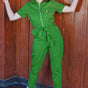 A person wearing a green boilersuit with a half sleeve shirt upper, straight legs, and an embroidered flower posy on the bust pocket, standing in front of a wooden background.