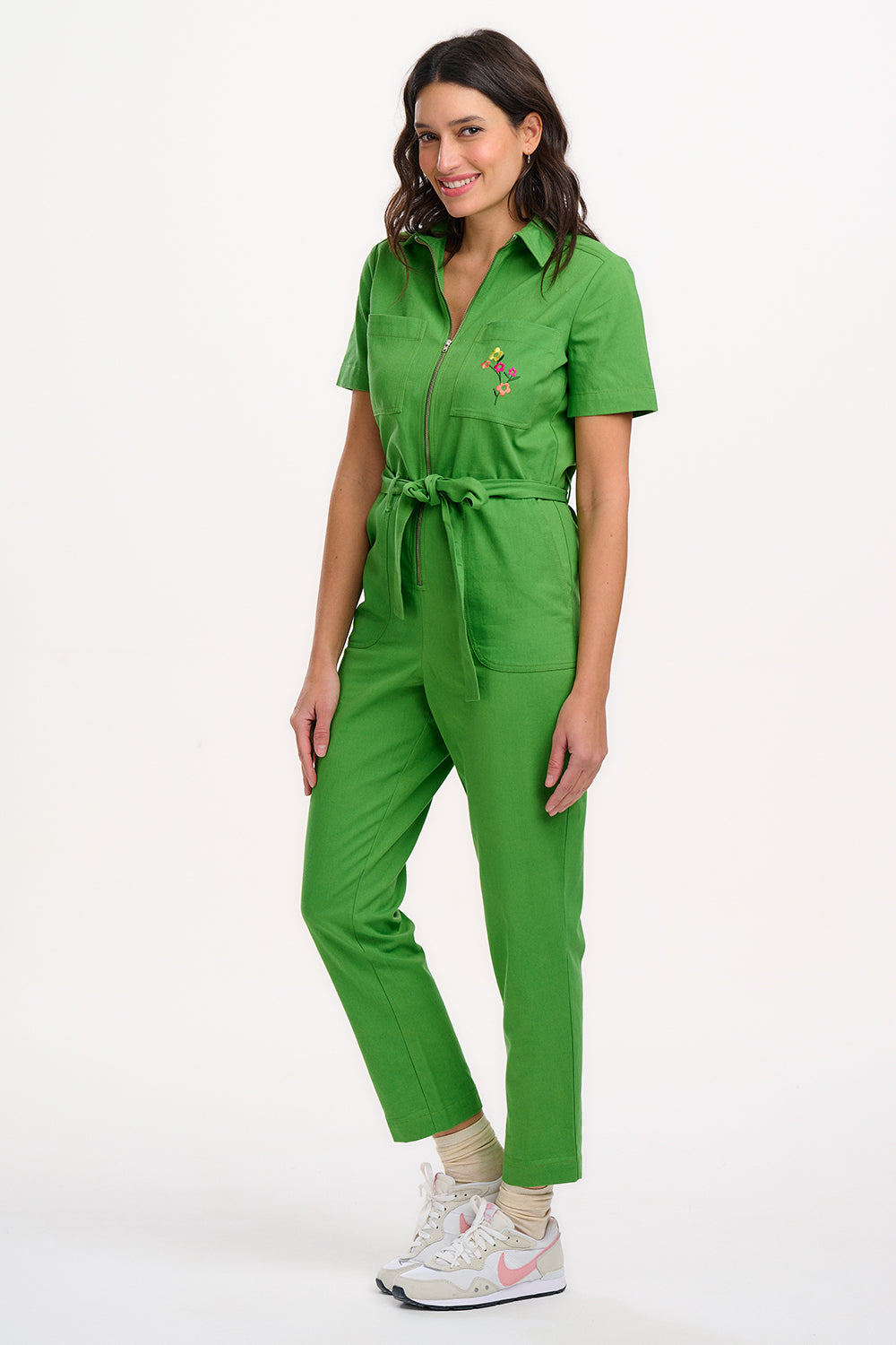 A model wearing a green boilersuit with rainbow flower embroidery on the chest pocket, featuring a half sleeve shirt upper, straight legs, and an exposed front zip.
