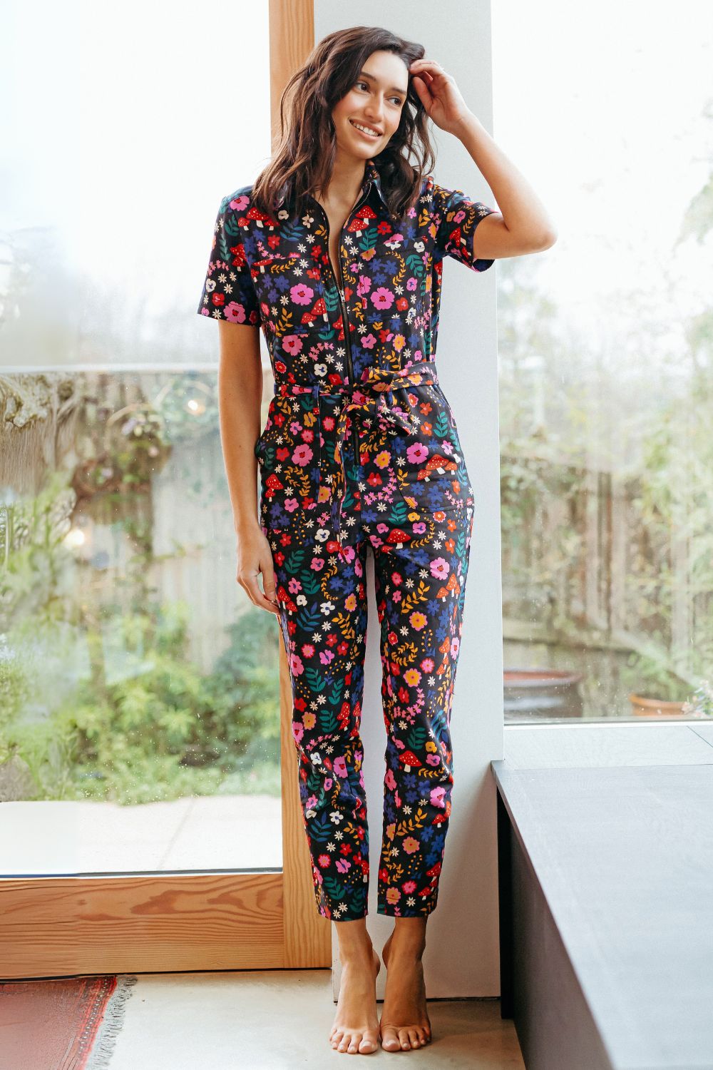 Sugarhill Brighton model wearing a floral jumpsuit, standing in a room with large windows.