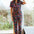 Sugarhill Brighton model wearing a floral jumpsuit, standing in a room with large windows.