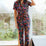 Sugarhill Brighton model wearing a floral jumpsuit, standing in a room with large windows.