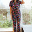 Sugarhill Brighton model wearing a floral jumpsuit, standing in a room with large windows.