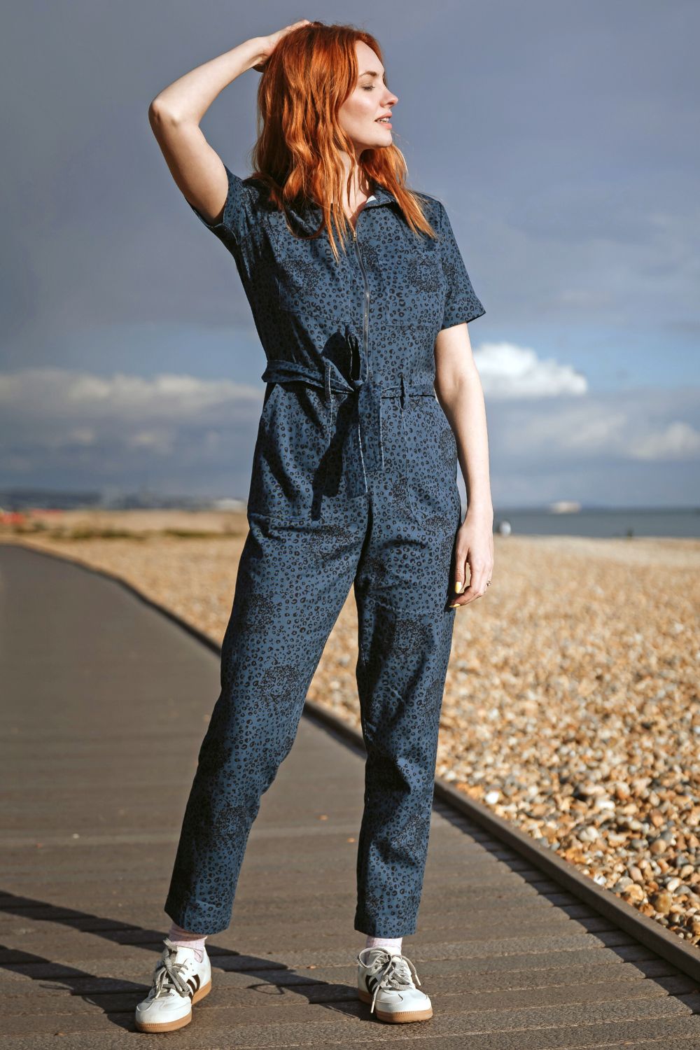 Woman in a blue patterned jumpsuit standing on a pebble beach with a cloudy sky.