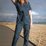 Woman in a blue patterned jumpsuit standing on a pebble beach with a cloudy sky.
