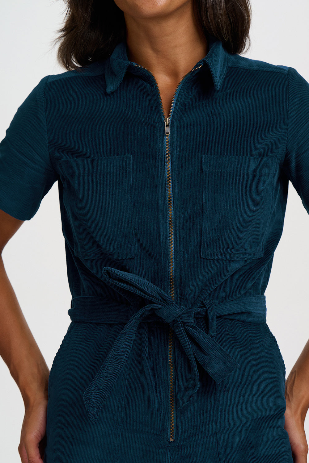 A close-up of a model wearing a dark teal corduroy shirt dress with a front zipper, chest pockets, and a tie belt at the waist.