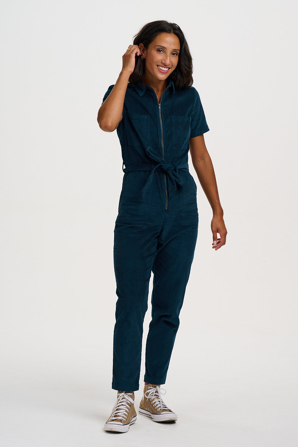 A woman modeling a dark teal corduroy jumpsuit with a front zipper, short sleeves, and a tie waist, paired with beige sneakers, against a plain white studio background.