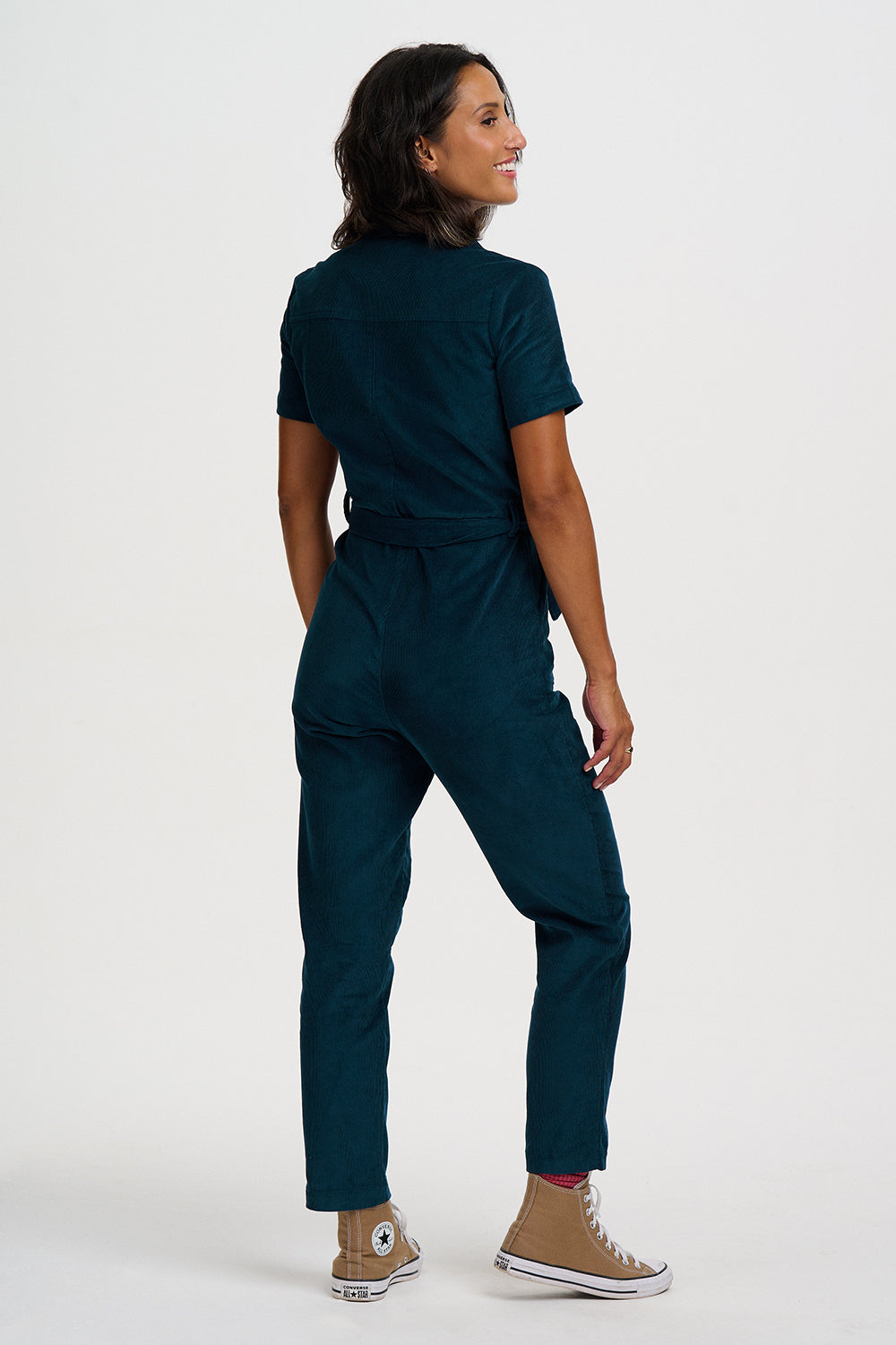 A woman modeling a dark teal short-sleeve scrub jumpsuit from the back, paired with tan high-top sneakers, against a plain white studio background.