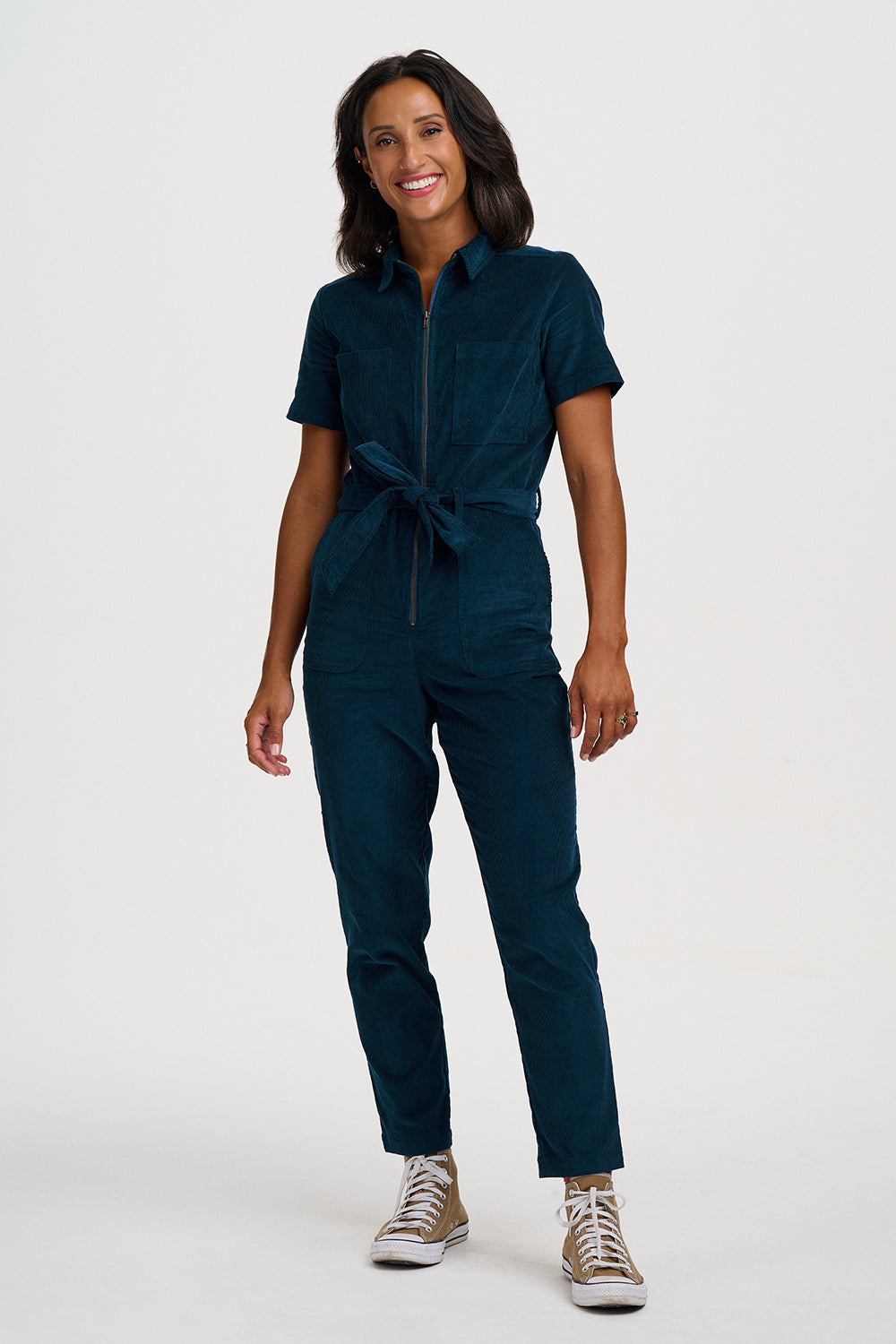 A woman modeling a dark teal short-sleeve jumpsuit with a front zipper and matching waist tie, paired with brown and white sneakers, against a plain white studio background.