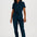 A woman modeling a dark teal short-sleeve jumpsuit with a front zipper and matching waist tie, paired with brown and white sneakers, against a plain white studio background.