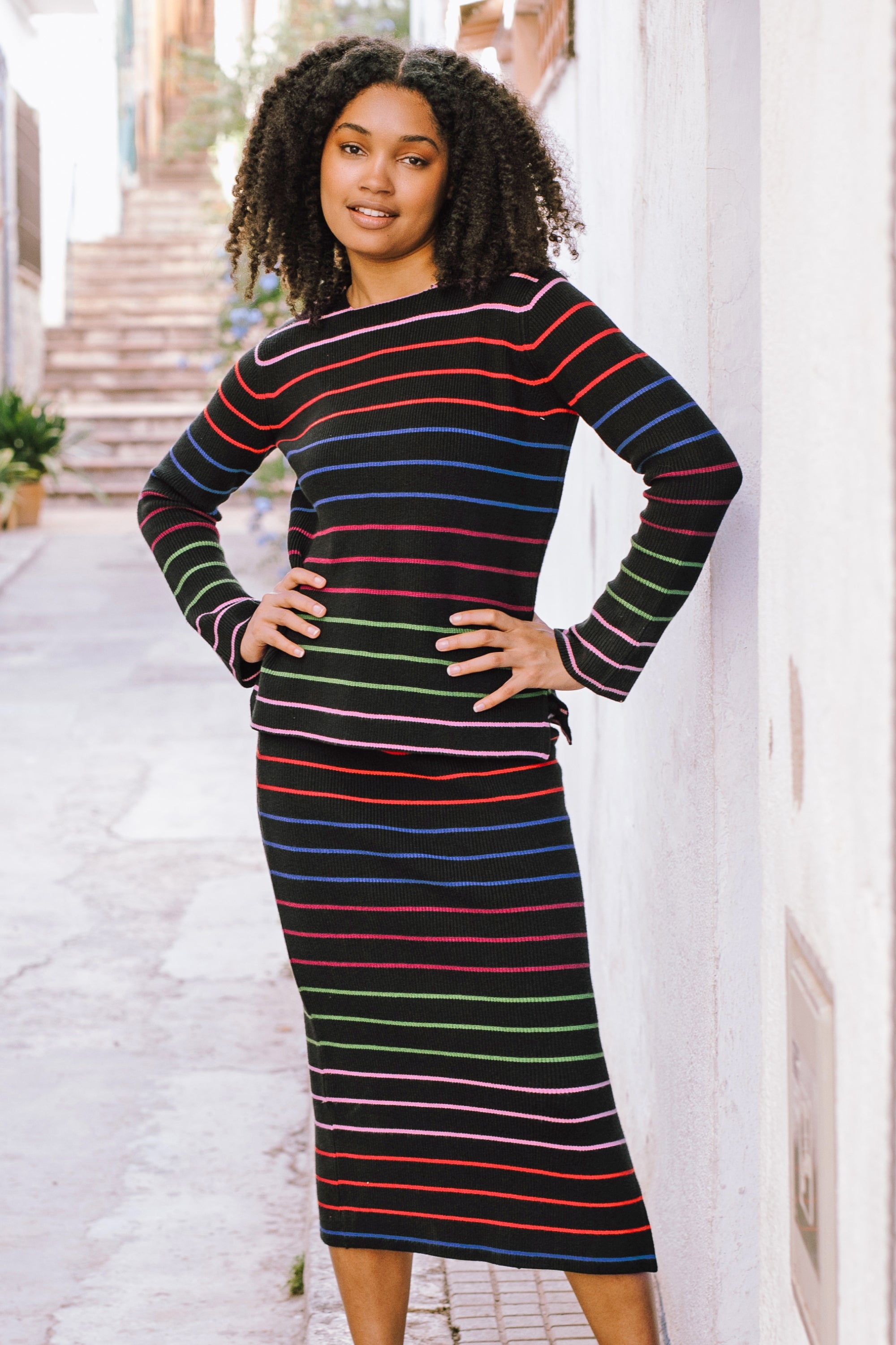 A model wearing a midi-length black knit skirt with thin rainbow stripes and an elastic waistband, paired with a long-sleeve top.