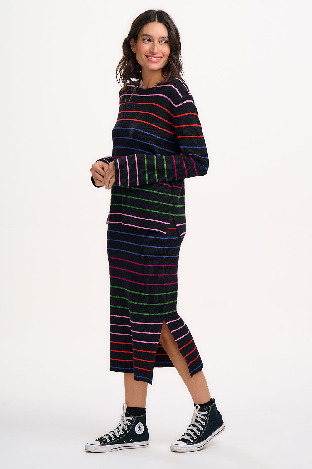 A model wearing a midi-length black knit skirt with thin rainbow stripes and an elastic waistband, paired with a long-sleeve top.