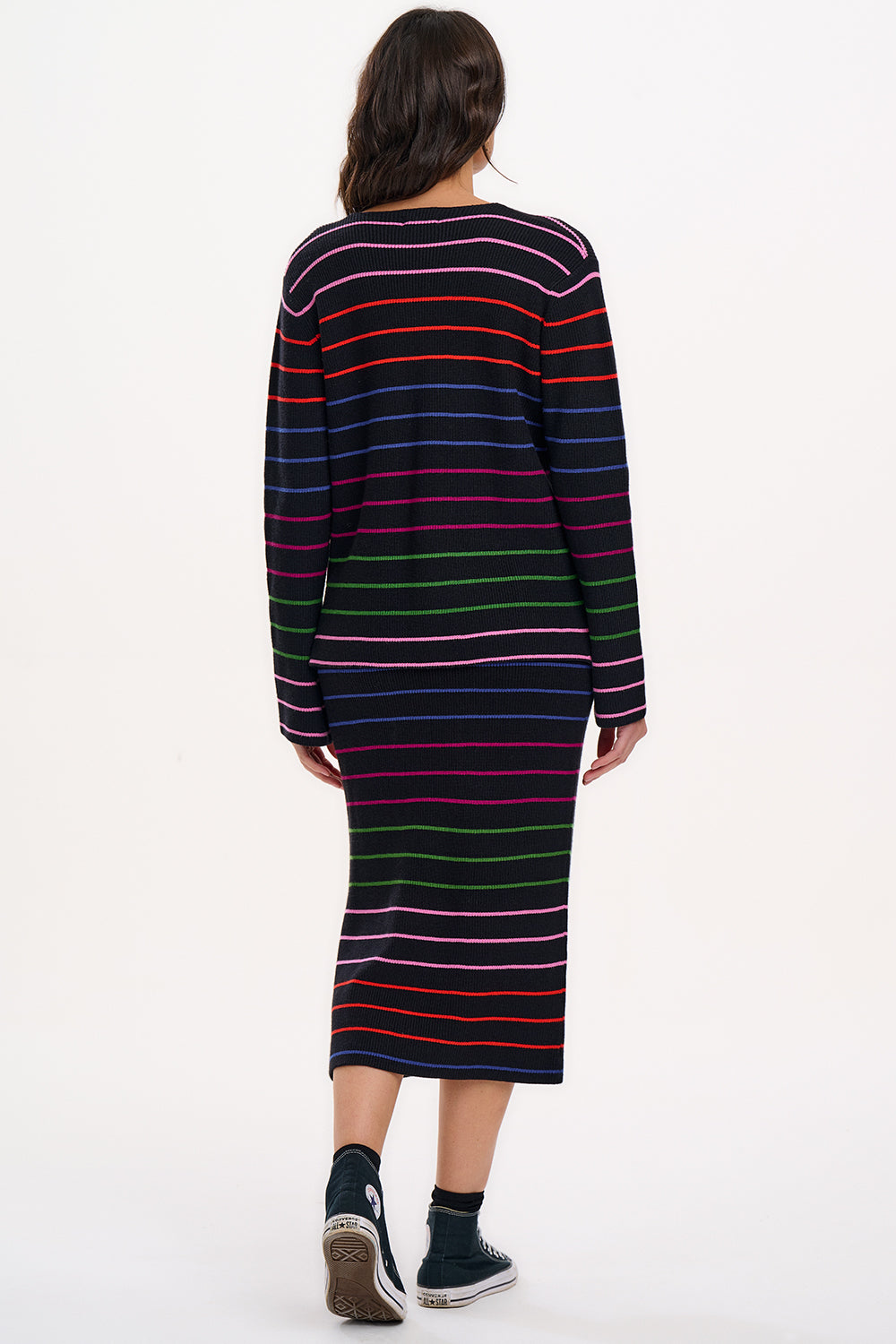 A model wearing a midi-length black knit skirt with thin rainbow stripes and an elastic waistband, paired with a long-sleeve top.