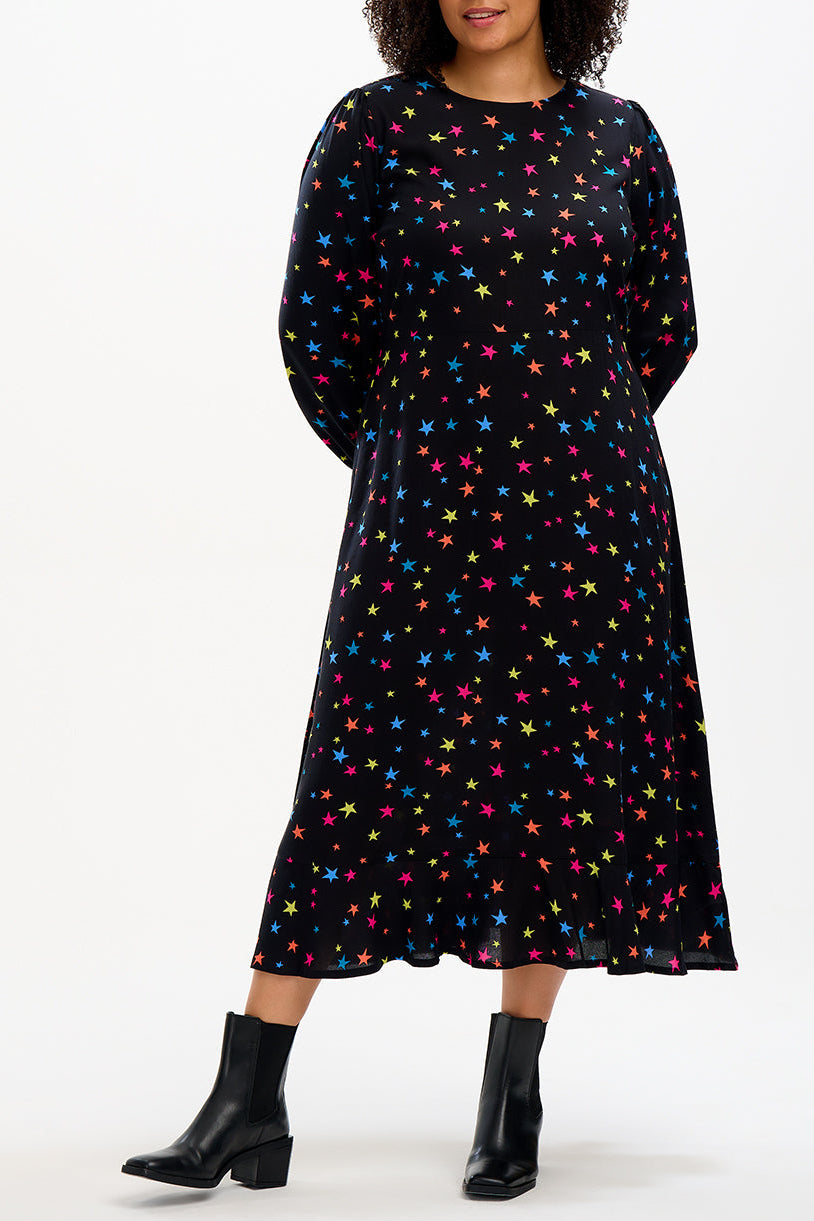 A woman standing and posing in a black midi dress with a rainbow star confetti pattern. 
