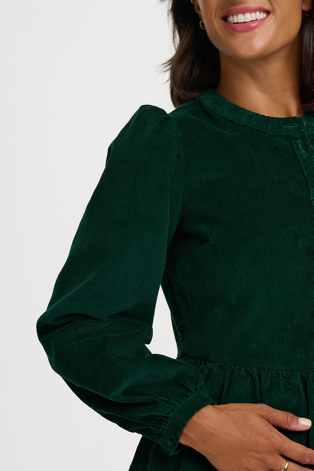 A close-up of a woman wearing a dark forest green corduroy long-sleeve dress with a gathered waist, against a plain light gray background.