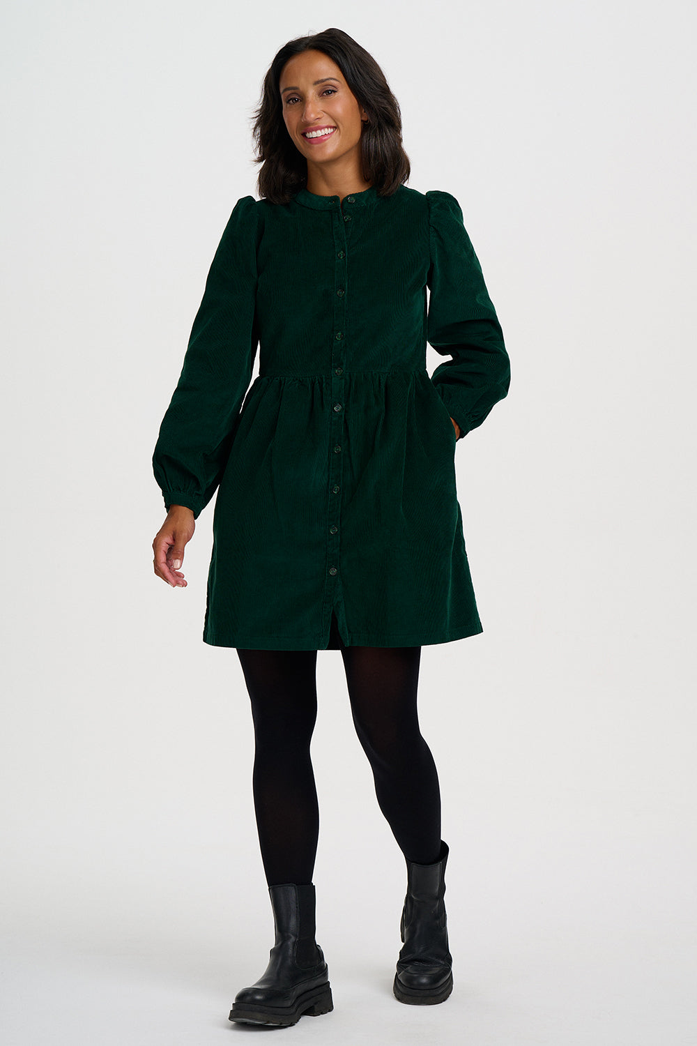 A woman modeling a dark green corduroy mini dress with long puff sleeves and a buttoned neckline, paired with black tights and black ankle boots, against a light gray studio background.