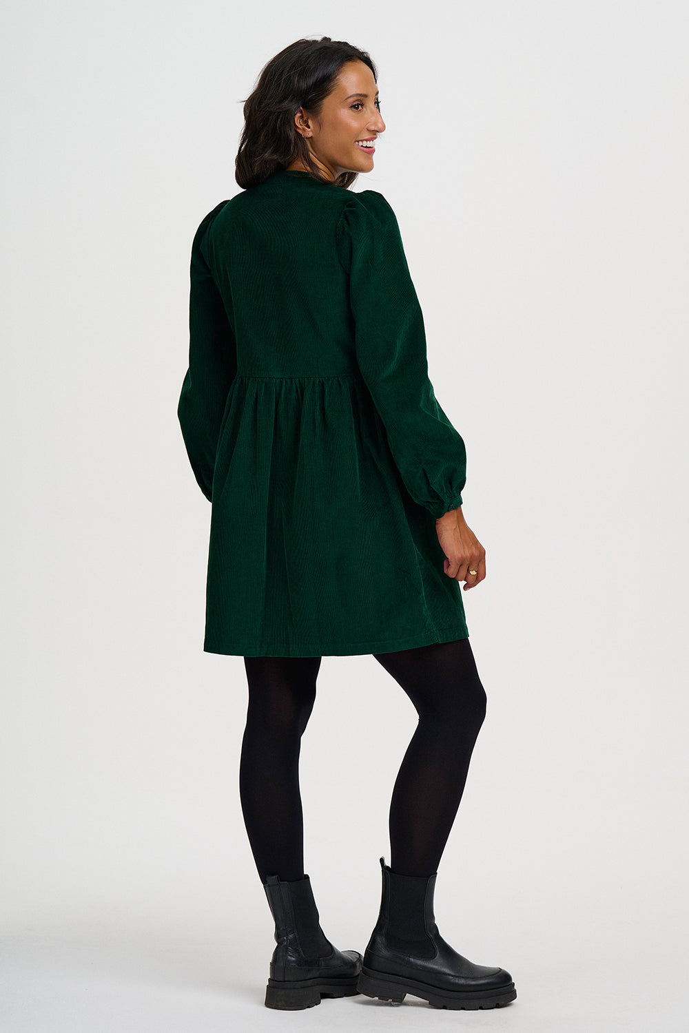 A woman modeling a dark green long-sleeve dress with a gathered waist, paired with black tights and black Chelsea boots, photographed against a plain light gray studio background.