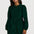 A woman modeling a dark green corduroy mini dress with long puff sleeves and a button-up front, styled with black tights and black ankle boots against a light gray studio background.