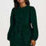 A woman modeling a dark green corduroy mini dress with long puff sleeves and a button-up front, styled with black tights and black ankle boots against a light gray studio background.