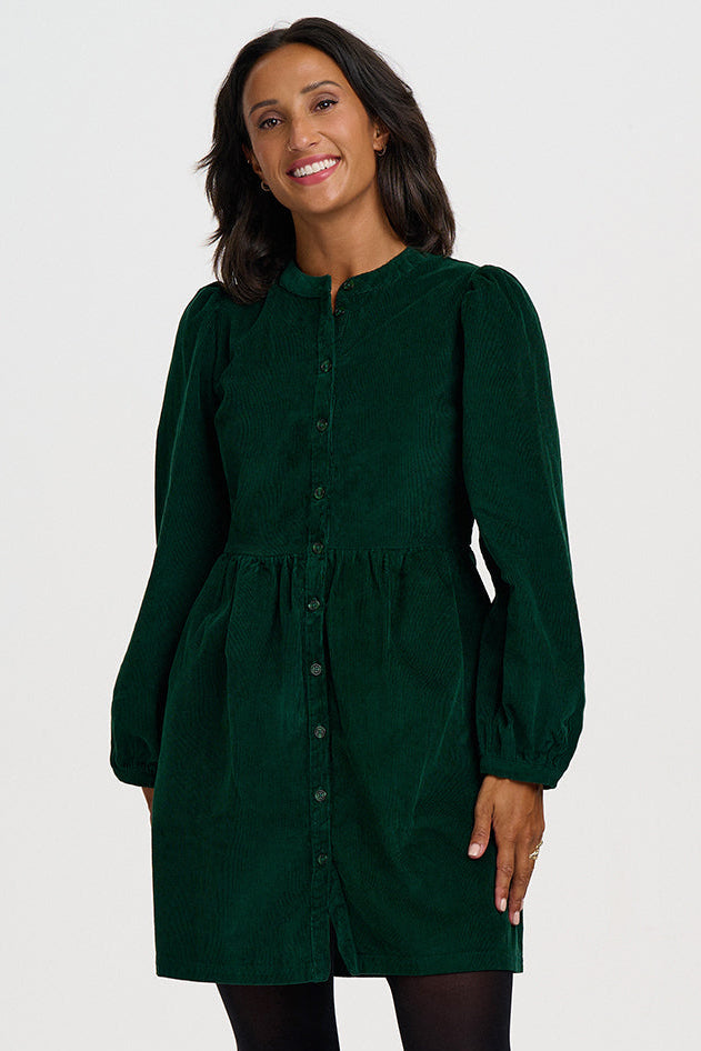 A woman modeling a dark green corduroy mini dress with long puff sleeves and a button-up front, styled with black tights and black ankle boots against a light gray studio background.