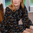 A woman with strawberry-blonde hair sits on a wooden chair wearing a black long-sleeve dress with a dense white and orange floral print.