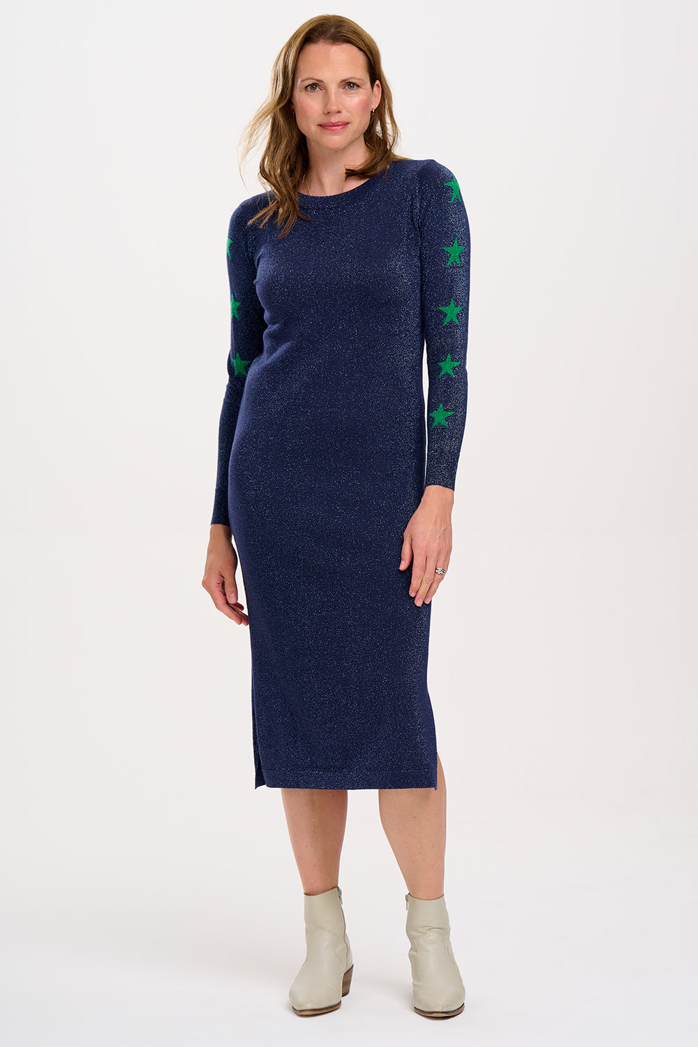 A woman standing and posing in a navy knit midi dress with star sleeves and a slit at the bottom. She is wearing white ankle boots.