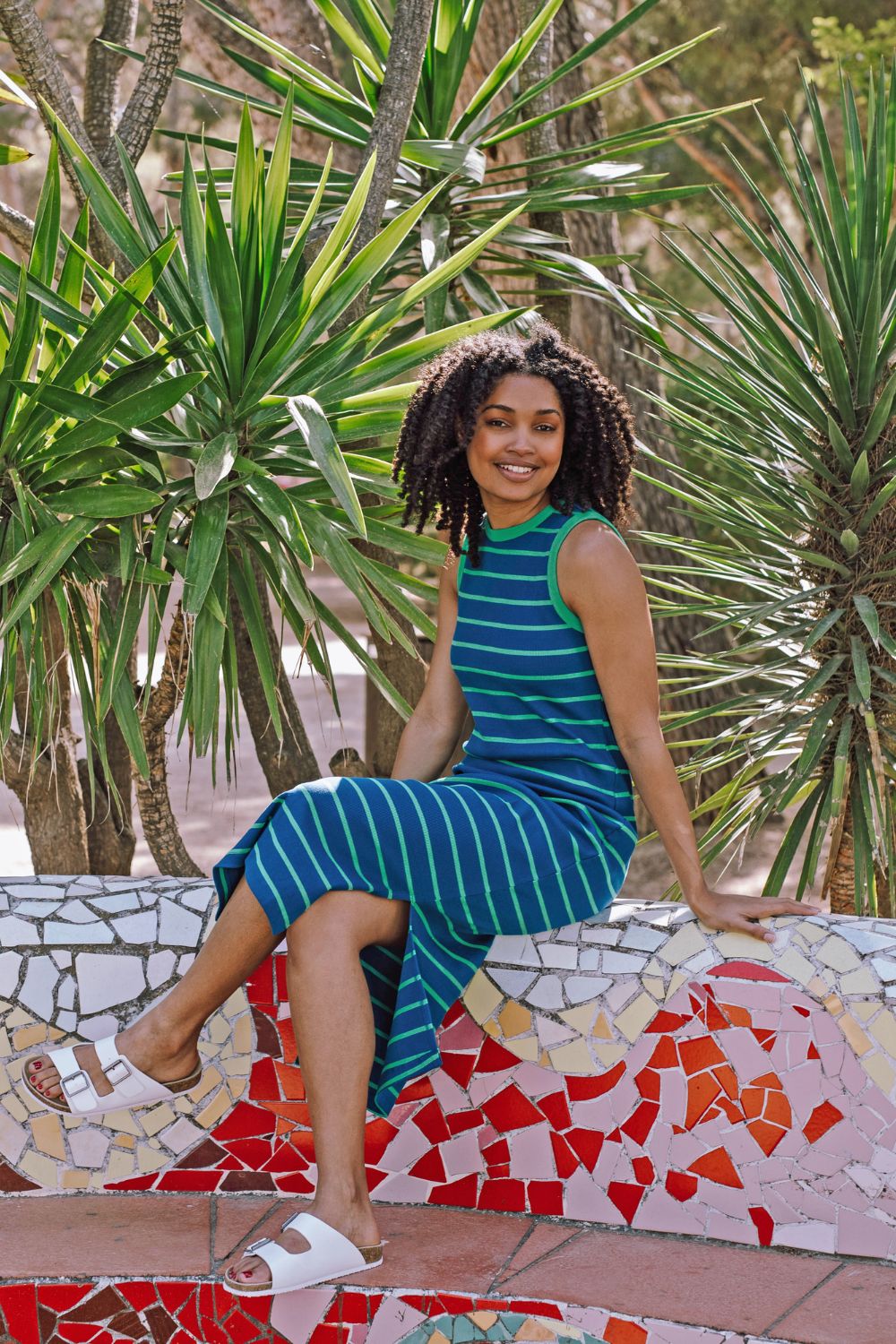 Woman in a blue dress sitting on a mosaic bench with green plants in the background