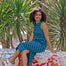Woman in a blue dress sitting on a mosaic bench with green plants in the background