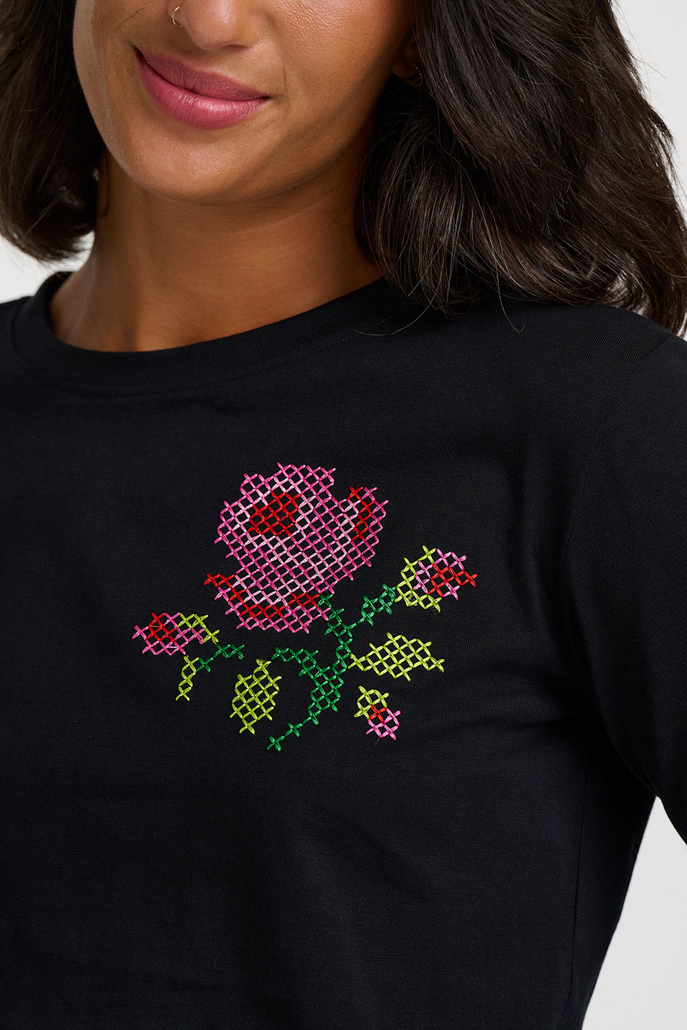 A close-up of a woman wearing a black crewneck t-shirt with a colorful cross-stitch style floral graphic on the chest.