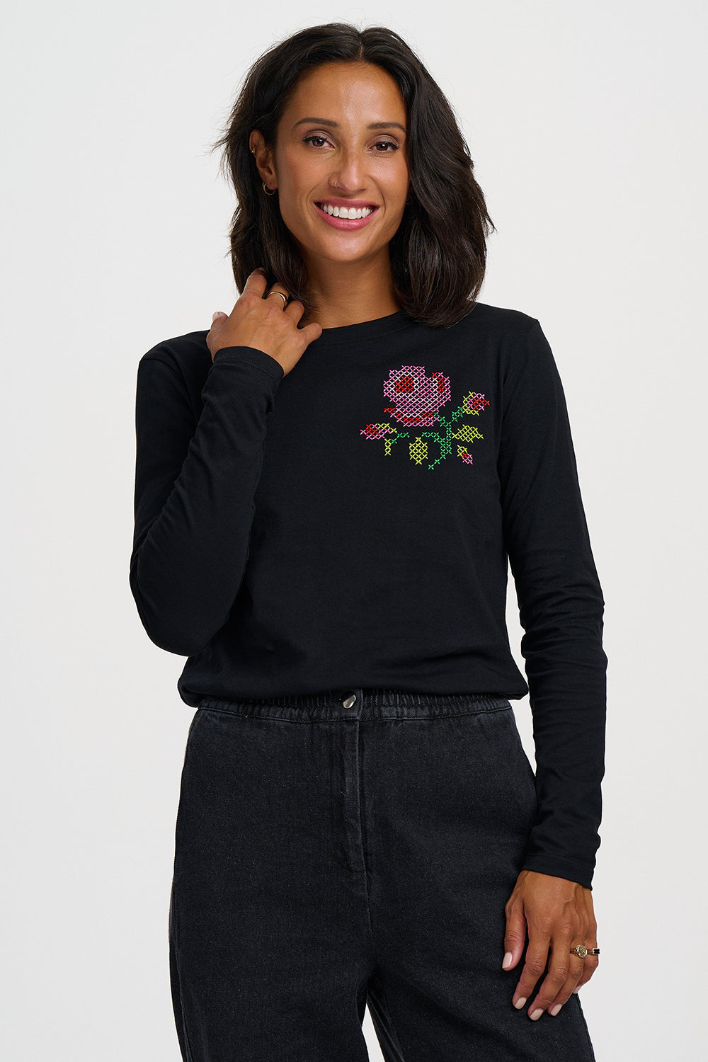 A woman wearing a black long-sleeve crewneck sweatshirt with a colorful embroidered rose on the left chest, paired with black jeans, against a light gray studio background.