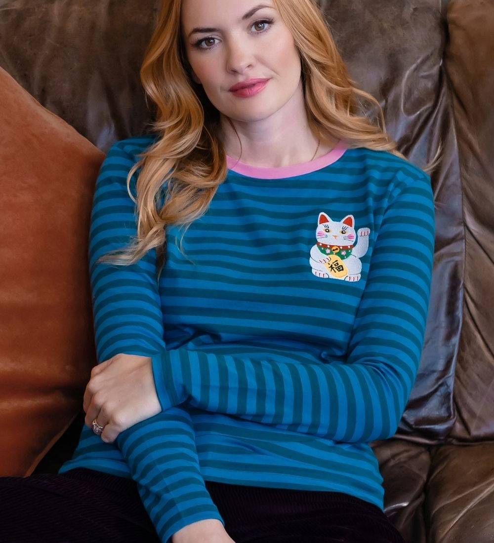 A woman with blonde hair sits on a brown leather couch wearing a teal and blue striped long-sleeve shirt with a pink collar and a white maneki-neko patch on the chest.