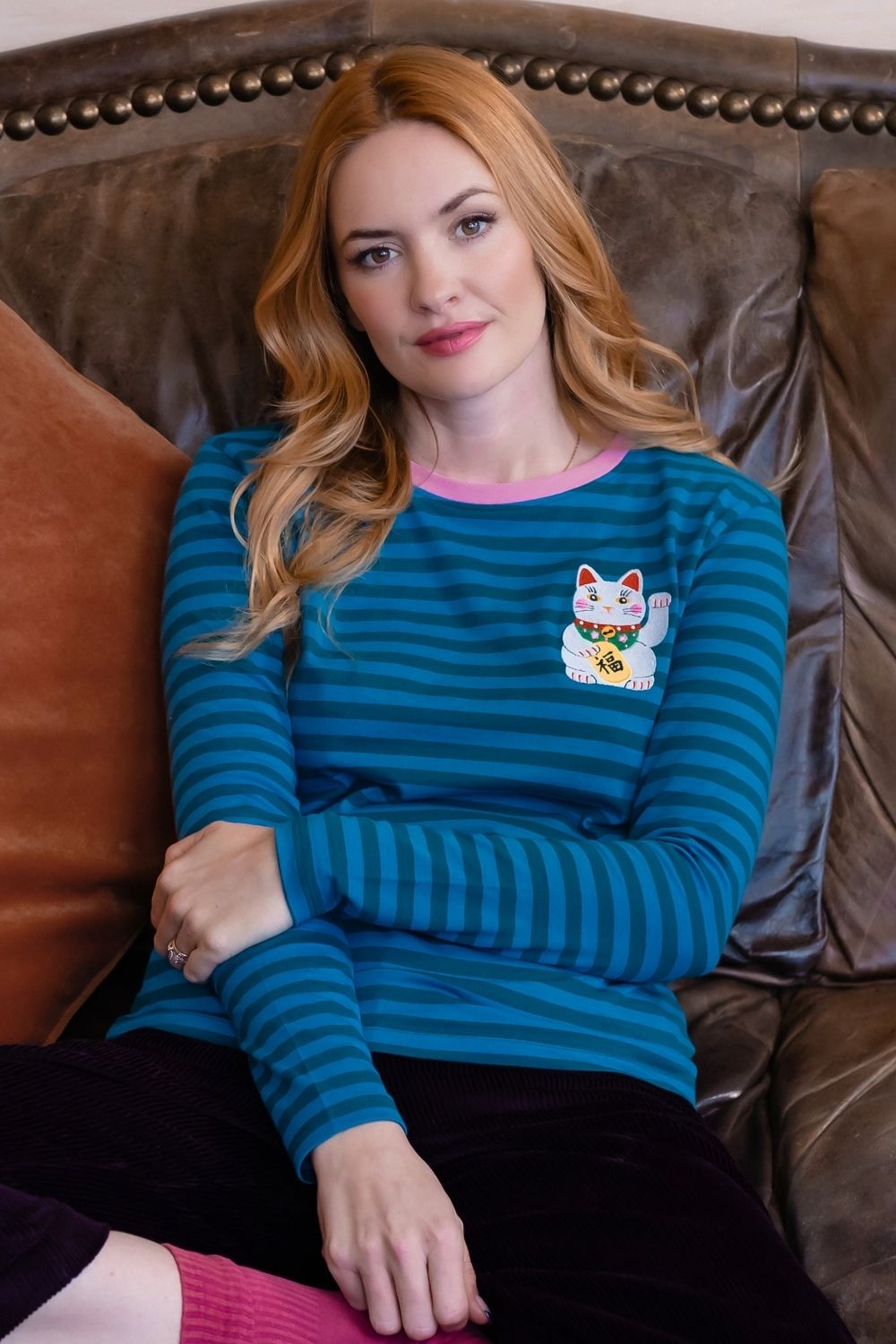 A woman with blonde hair sits on a brown leather couch wearing a teal and blue striped long-sleeve shirt with a pink collar and a white maneki-neko patch on the chest.