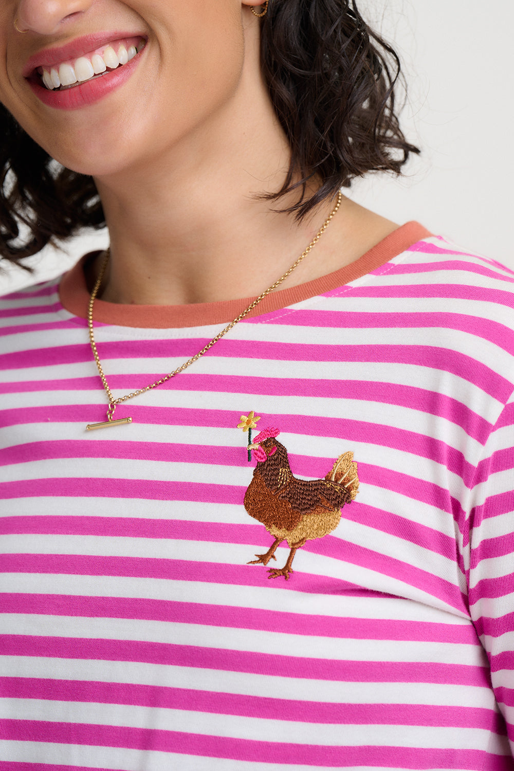 A close-up of a person wearing a pink and white striped t-shirt with a gold embroidered chicken on the chest, paired with a gold necklace.