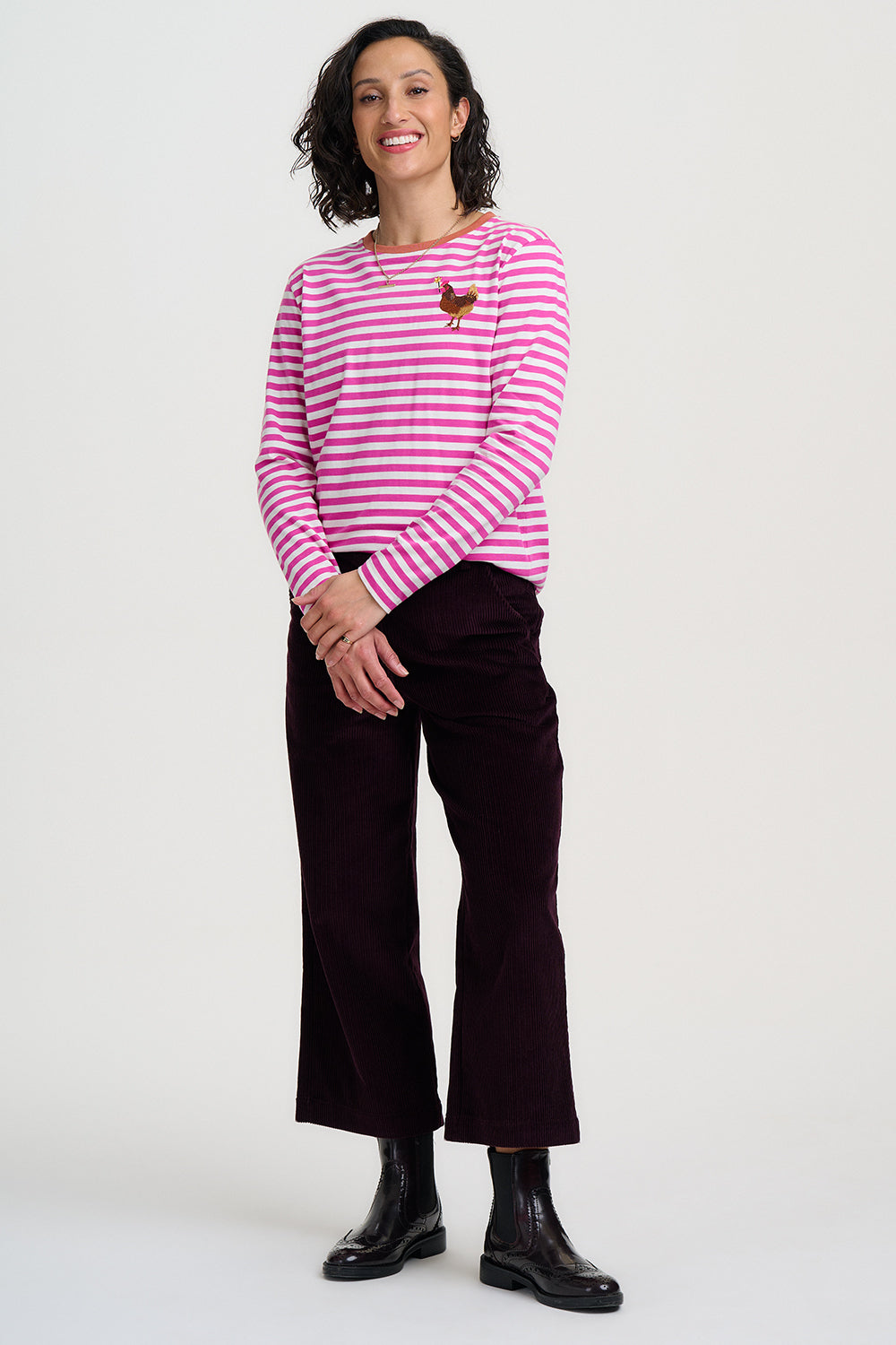 A woman modeling a pink and white striped long-sleeve top with a small embroidered rooster on the chest, paired with dark burgundy cropped trousers and black boots, against a plain light gray studio background.