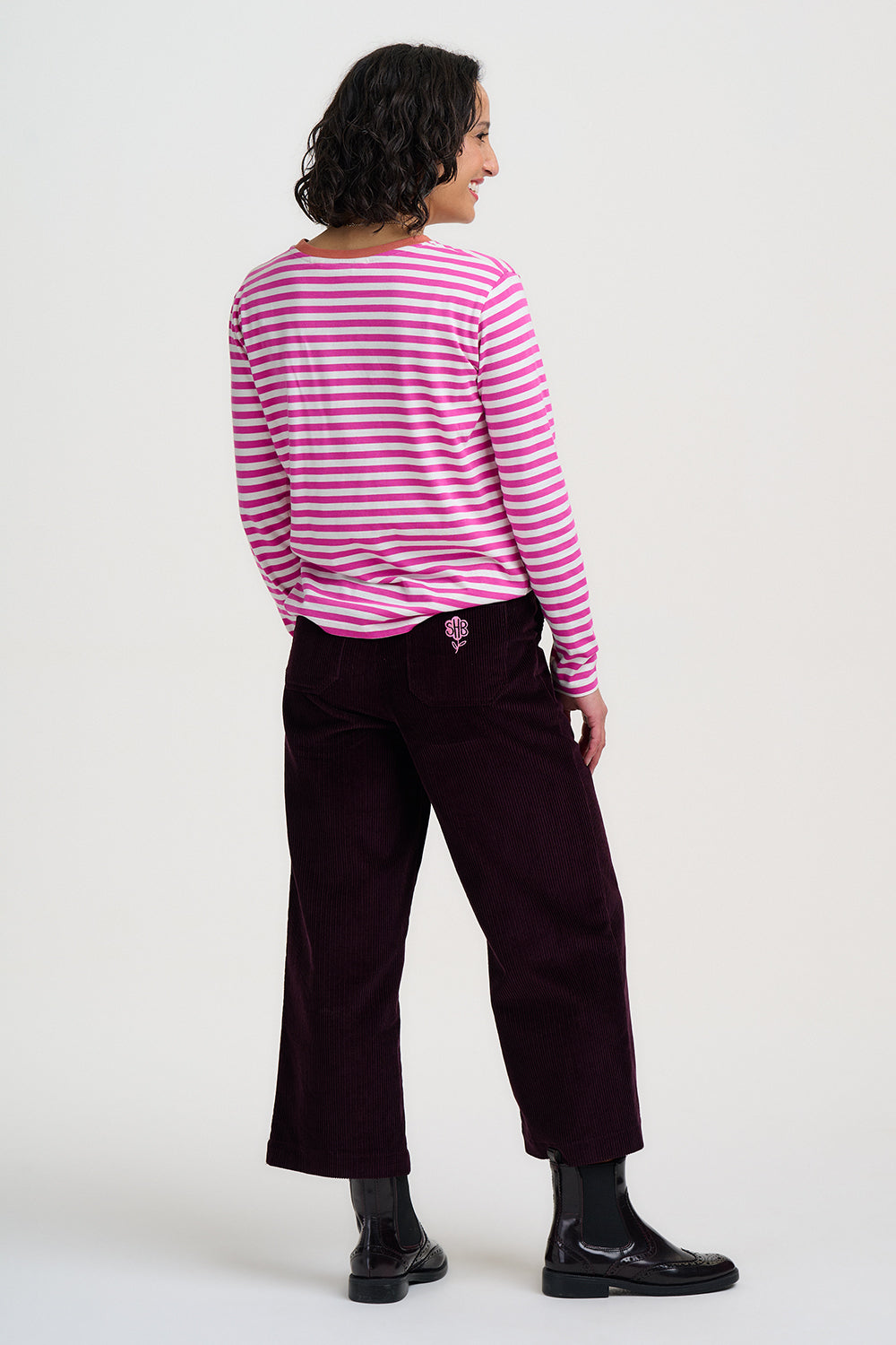 A woman shown from the back wearing a pink and white striped long-sleeve top, dark burgundy cropped trousers, and black boots, photographed against a plain light gray studio background.