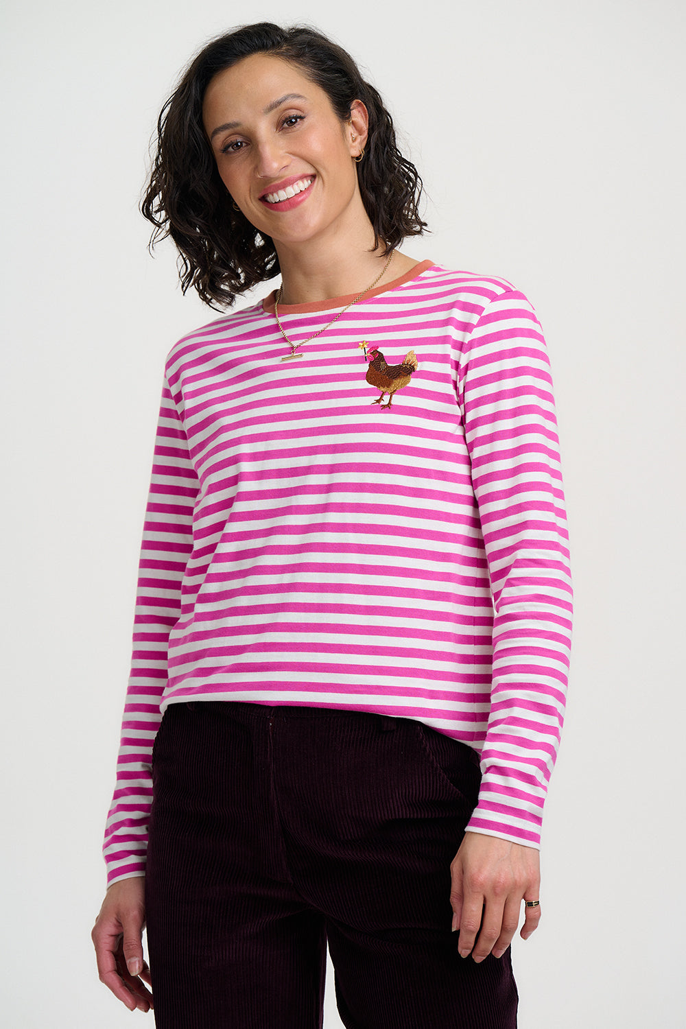 A woman wearing a pink and white striped long-sleeve shirt with a small embroidered chicken on the left chest, paired with dark burgundy pants, against a light gray studio background.