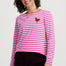 A woman wearing a pink and white striped long-sleeve shirt with a small embroidered chicken on the left chest, paired with dark burgundy pants, against a light gray studio background.