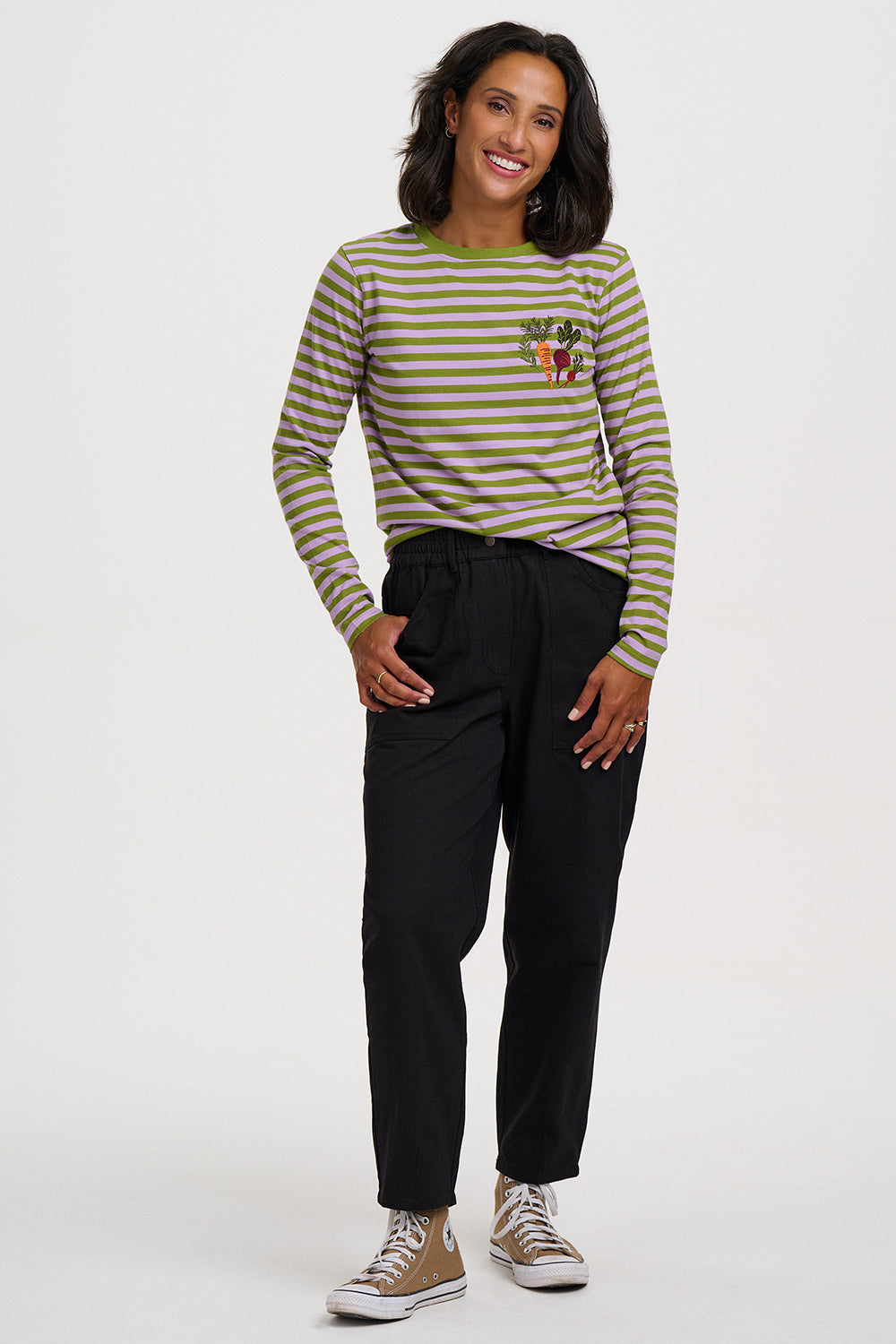 A woman modeling a long-sleeve striped top with olive green and white horizontal stripes, a small colorful chest graphic, black pants, and beige sneakers, photographed against a plain light gray studio background.