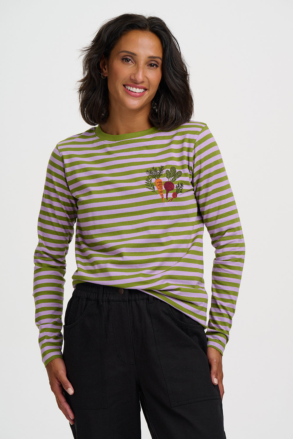 A woman wearing a long-sleeve striped top with olive green and light pink horizontal stripes, featuring a small embroidered vegetable motif on the left chest, paired with black pants, against a light gray studio background.