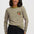 A woman wearing a long-sleeve striped top with olive green and light pink horizontal stripes, featuring a small embroidered vegetable motif on the left chest, paired with black pants, against a light gray studio background.