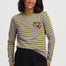 A woman wearing a long-sleeve striped top with olive green and light pink horizontal stripes, featuring a small embroidered vegetable motif on the left chest, paired with black pants, against a light gray studio background.