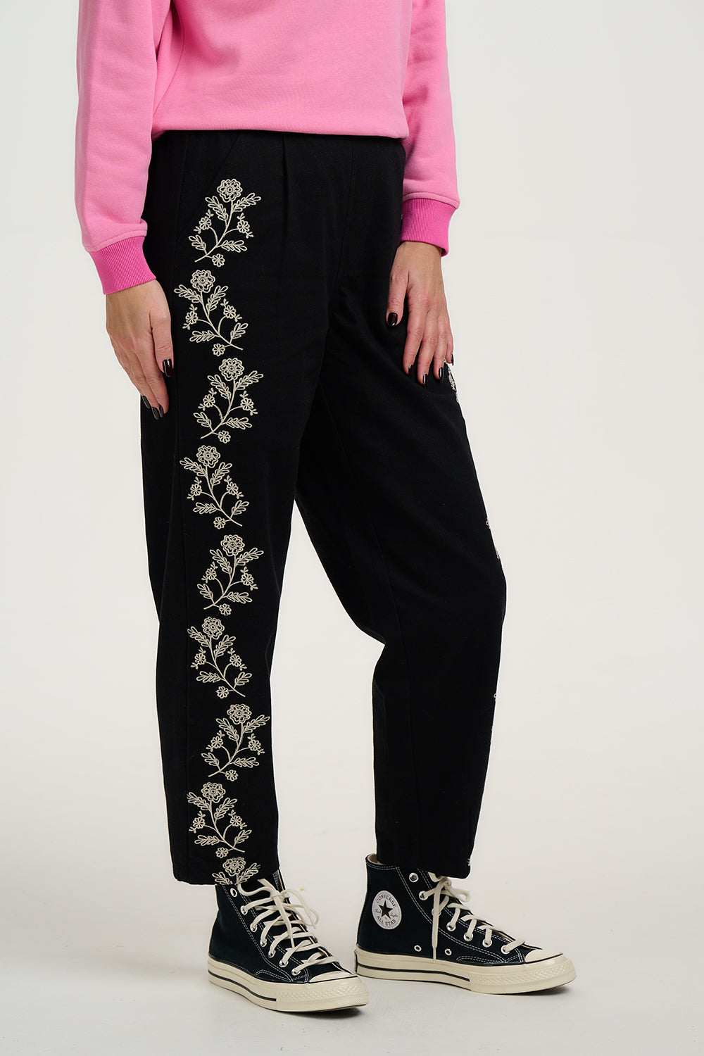 Person wearing black pants with floral embroidery and a pink sweatshirt on a white background
