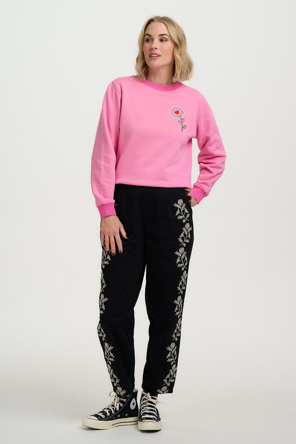 Woman wearing a pink sweatshirt with a floral design and black pants with white patterns on a white background