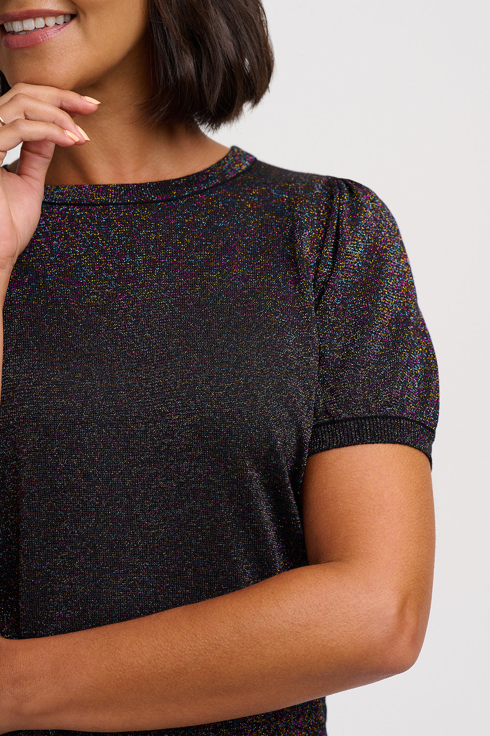 A close-up of a woman wearing a short-sleeve crewneck top with a black and multicolor micro-iridescent knit texture, set against a plain light gray background.