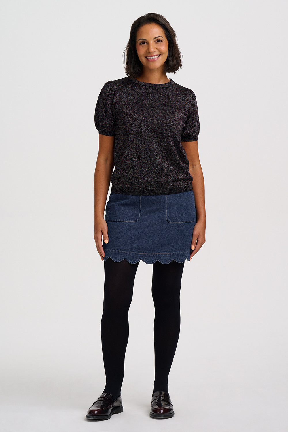 A woman modeling a short-sleeve black knit top, a dark blue denim mini skirt with a scalloped hem, black tights, and black leather shoes against a light gray studio background.