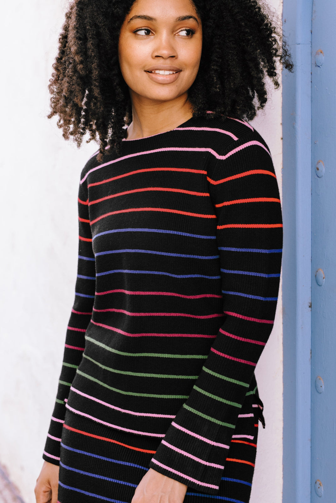 A woman wearing a black long-sleeved jumper with horizontal rainbow stripes in pink, red, blue, and green.