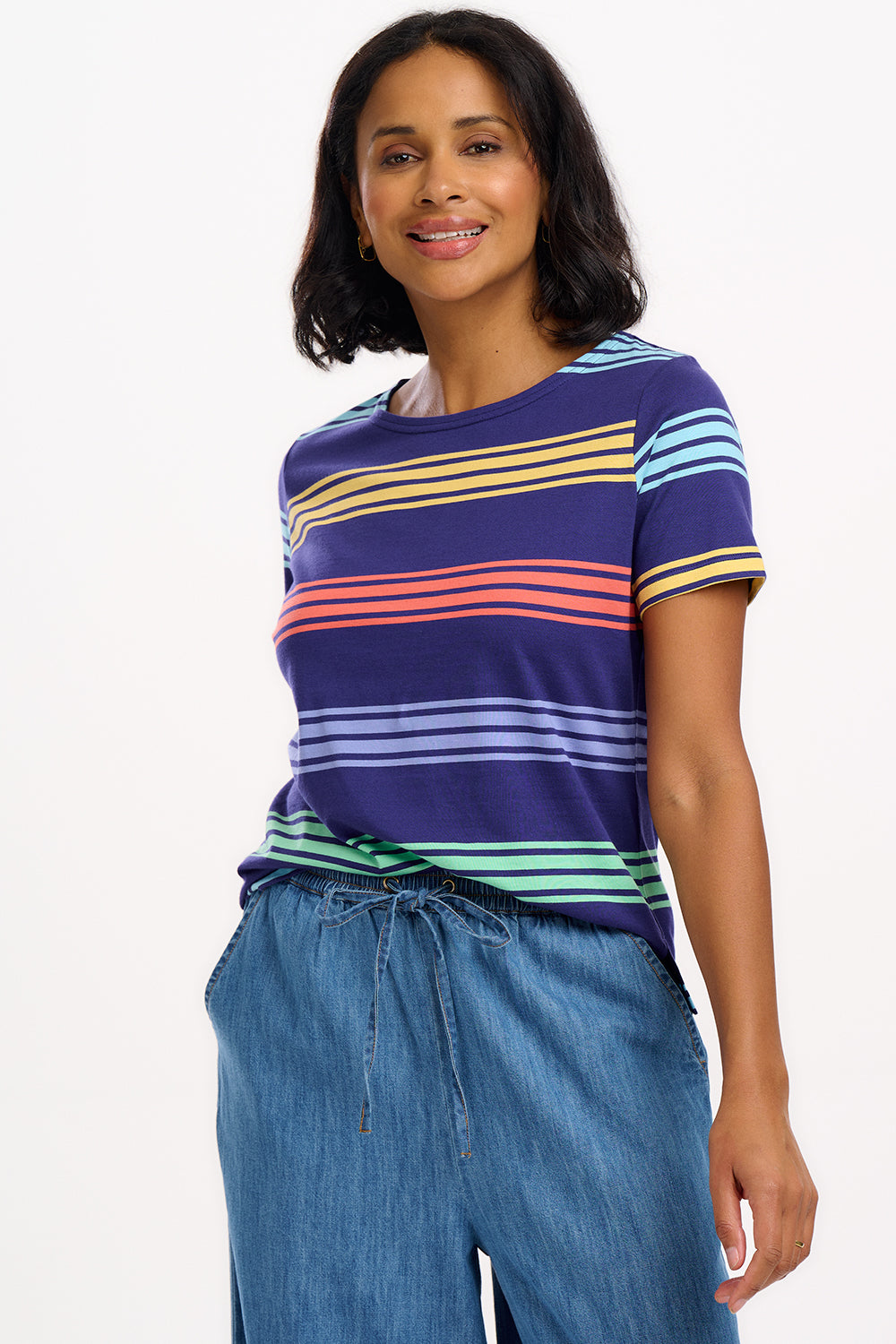 A woman wearing a navy t-shirt with wide muted stripes in mustard, coral, mint green, and baby blue over a navy base, paired with blue jeans.