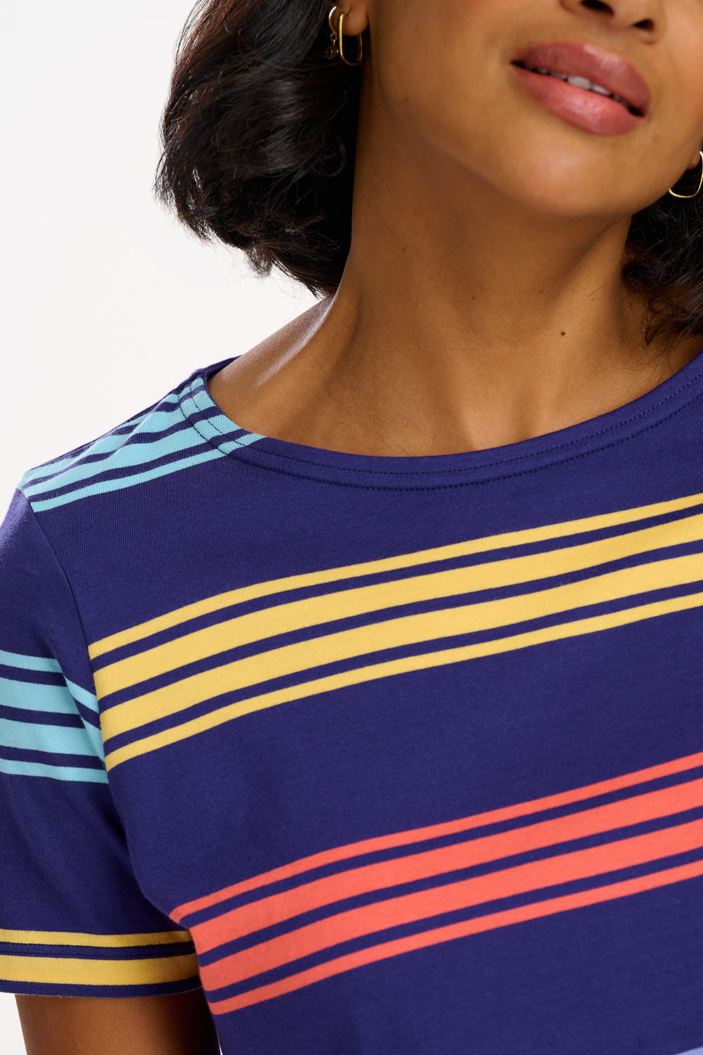A woman wearing a navy t-shirt with wide muted stripes in mustard, coral, mint green, and baby blue over a navy base, paired with blue jeans.